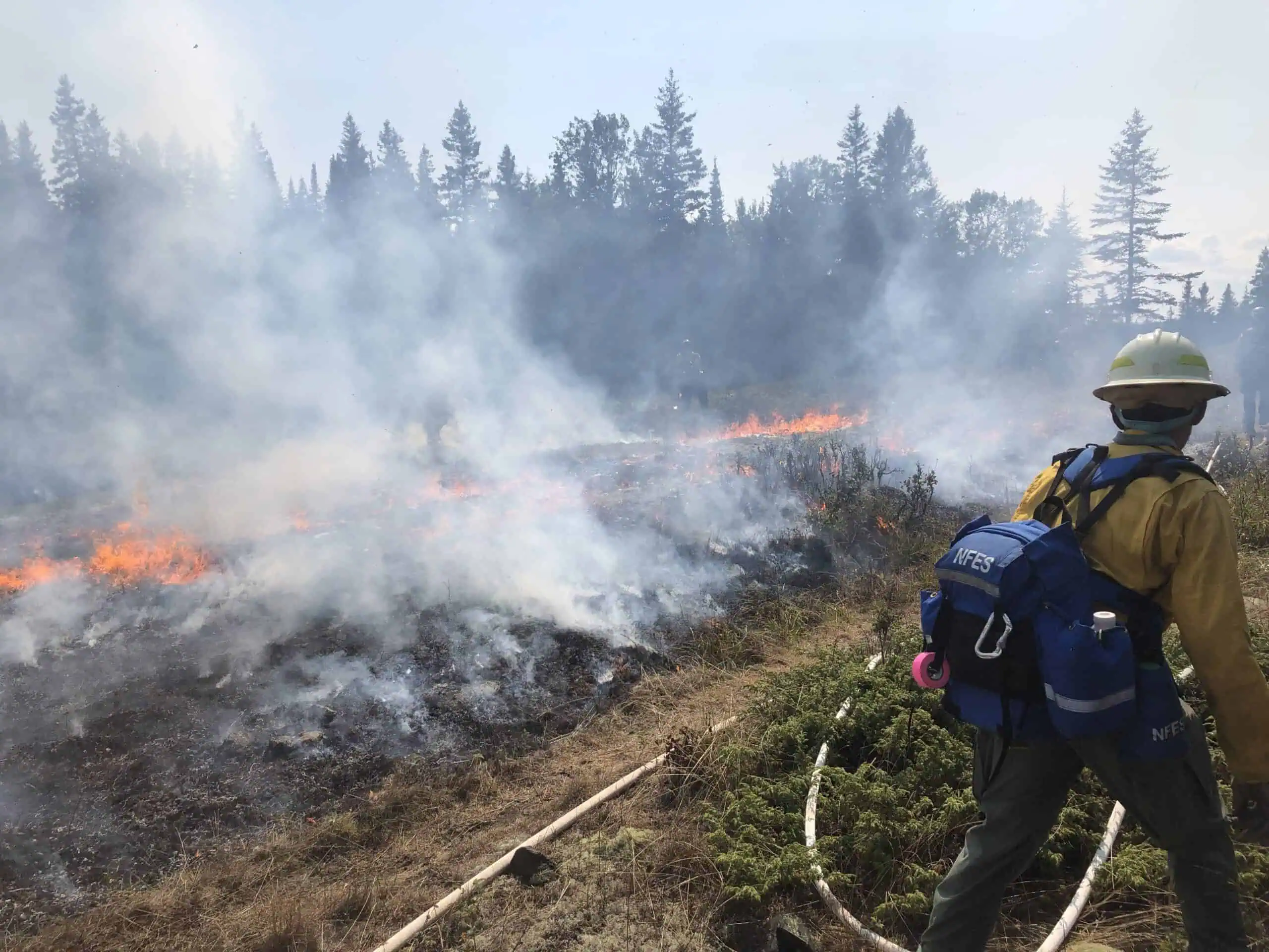 Wildland firefighter in yellow protective gear beside flames and smoke on an Isle Royale fireline.