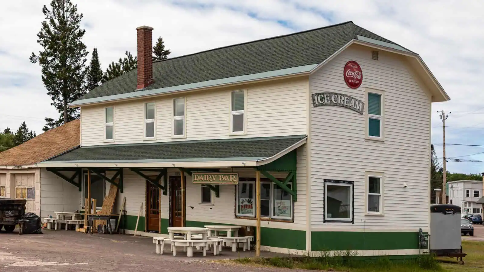 A two-storey cream and green clapboard building with a long covered platform canopy along its front, hung with ICE CREAM and Coca-Cola signs and a DAIRY BAR sign under the canopy, with picnic tables on the gravel lot outside.