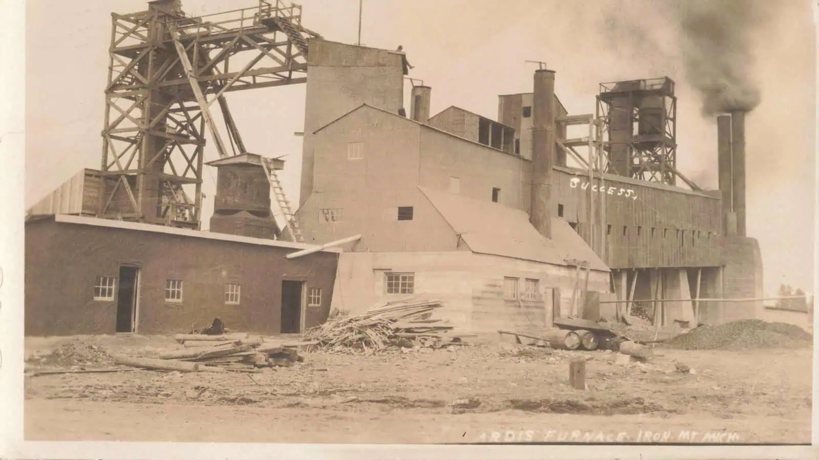 A sepia 1910 photo postcard of the Ardis Furnace plant in Iron Mountain, showing a timber headframe, a tall concrete building and a smoking stack, with the word SUCCESS hand-lettered in white on the building and the caption ARDIS FURNACE, IRON MT MICH lettered along the bottom edge.