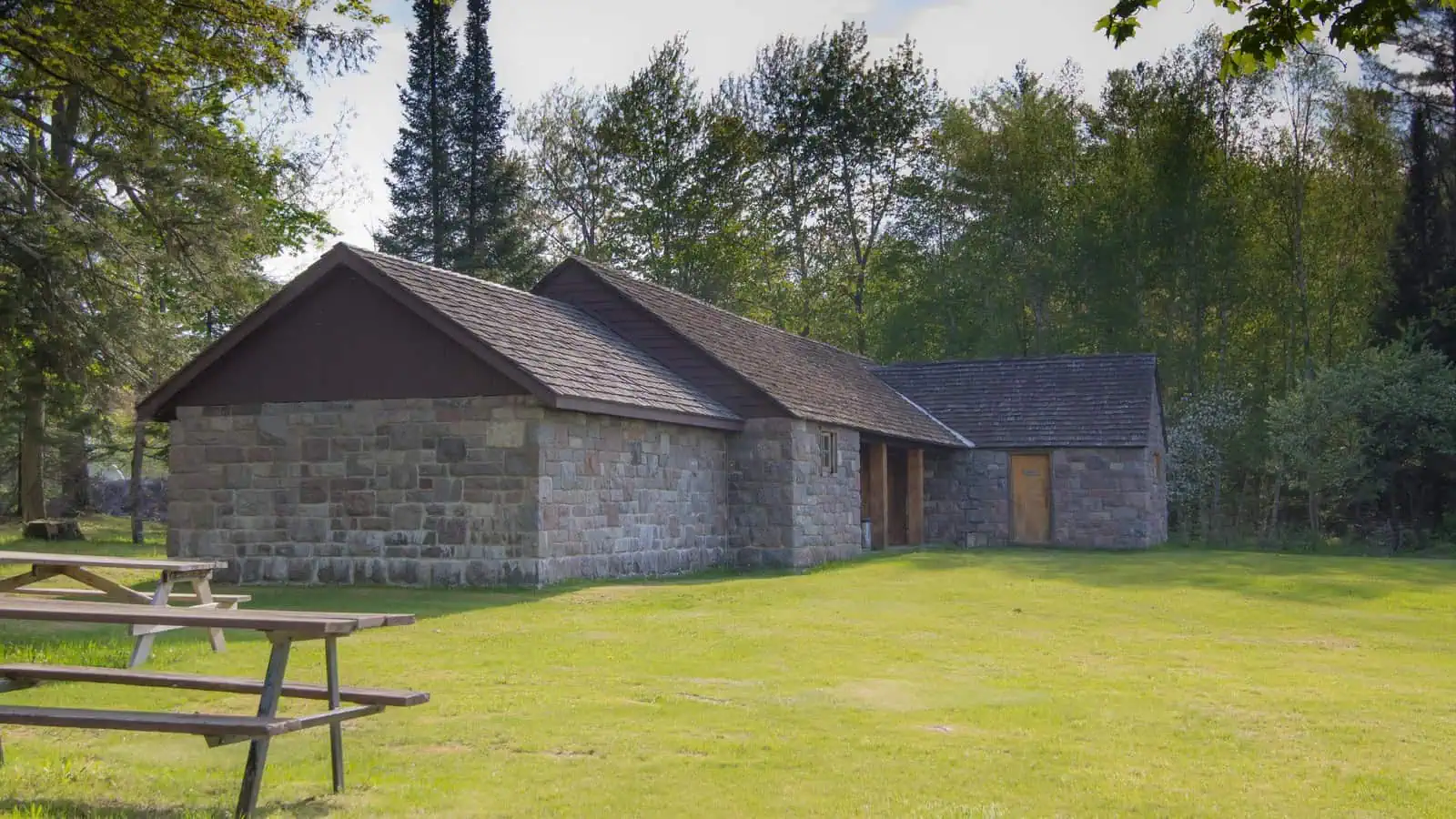 A low fieldstone picnic shelter with a shingled roof standing on mown grass among pines, with wooden picnic tables in the foreground.