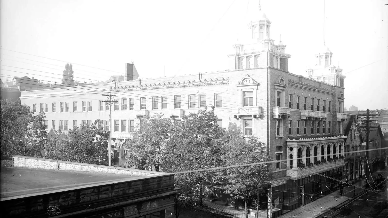 The Douglass House hotel in Houghton around 1908, a long light brick building with an arched ground-floor loggia and ornate corner cupolas, seen from above with streetcar tracks and trees along the street below.