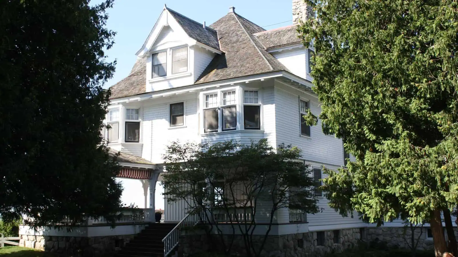 A large white three-story shingled cottage with a wide covered porch, dormer windows and a fieldstone foundation, framed by dark cedar trees, with a wooden stair leading up from a lawn edged with white flowers.