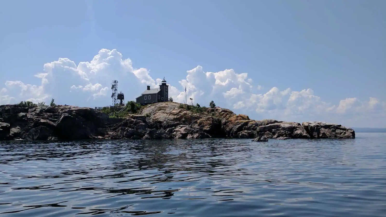 A low bare granite island in calm blue water with a small square stone lighthouse tower and attached house on its summit, a lattice mast beside them, and towering white clouds behind.