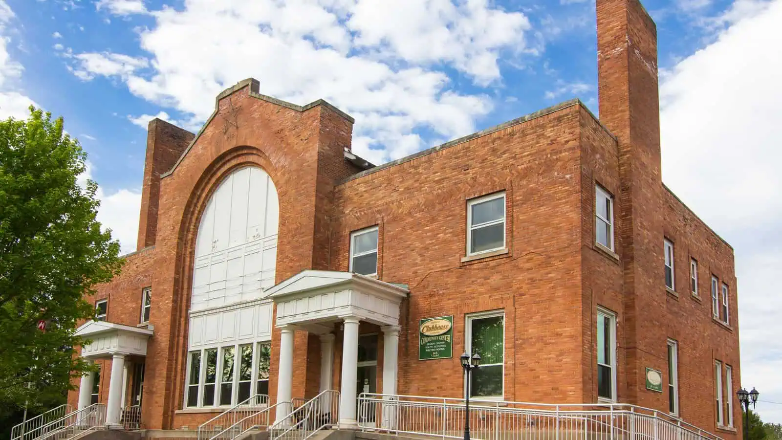 The Gwinn Community Clubhouse, a two-storey red brick building with a tall arched window, white columned entry porches and a brick chimney, standing on a lawn under a partly clouded sky.