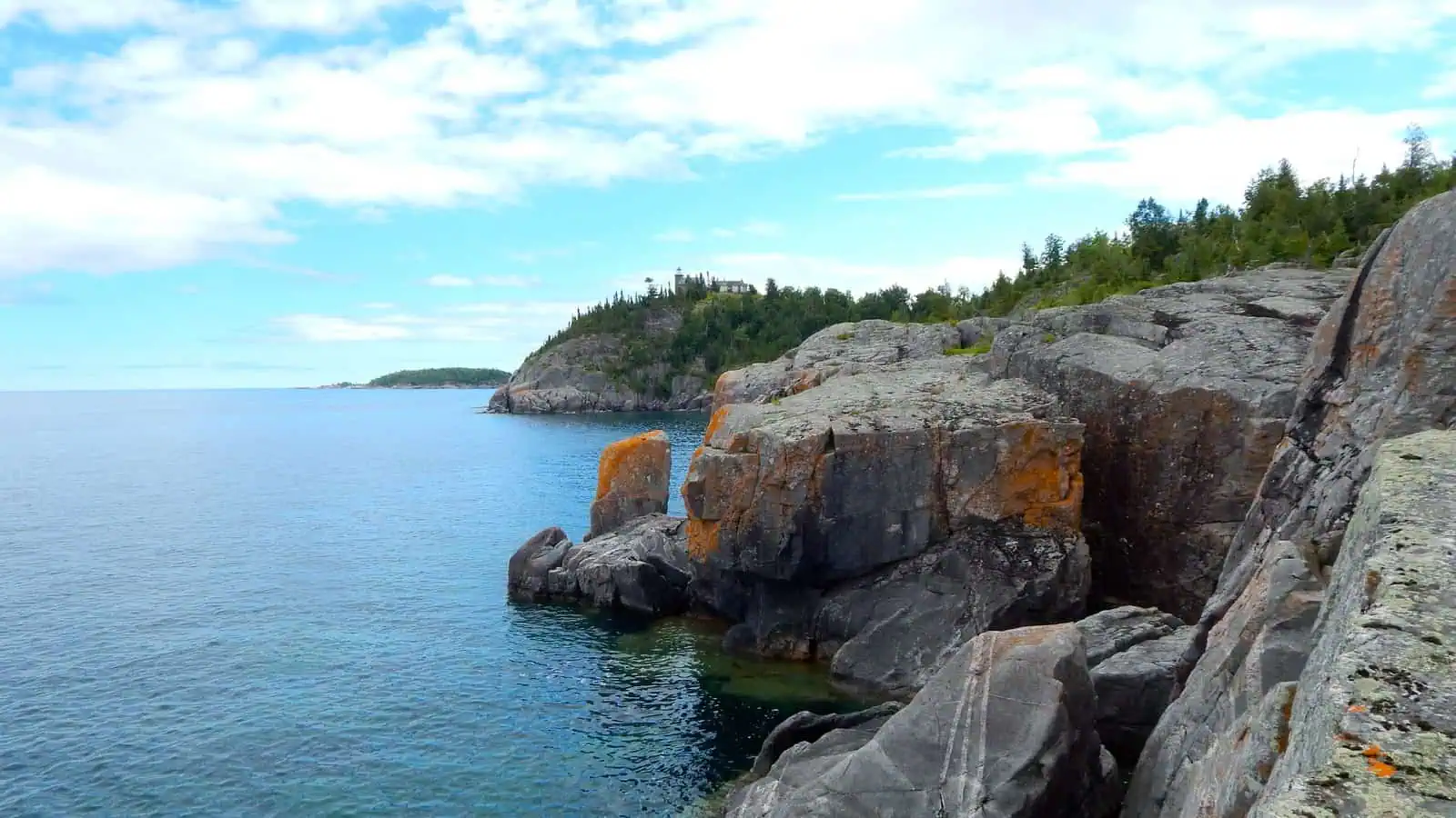 Grey granite ledges streaked with orange lichen drop into clear blue Lake Superior water, and on the wooded clifftop in the distance a small white lighthouse and keeper's house stand above the trees.