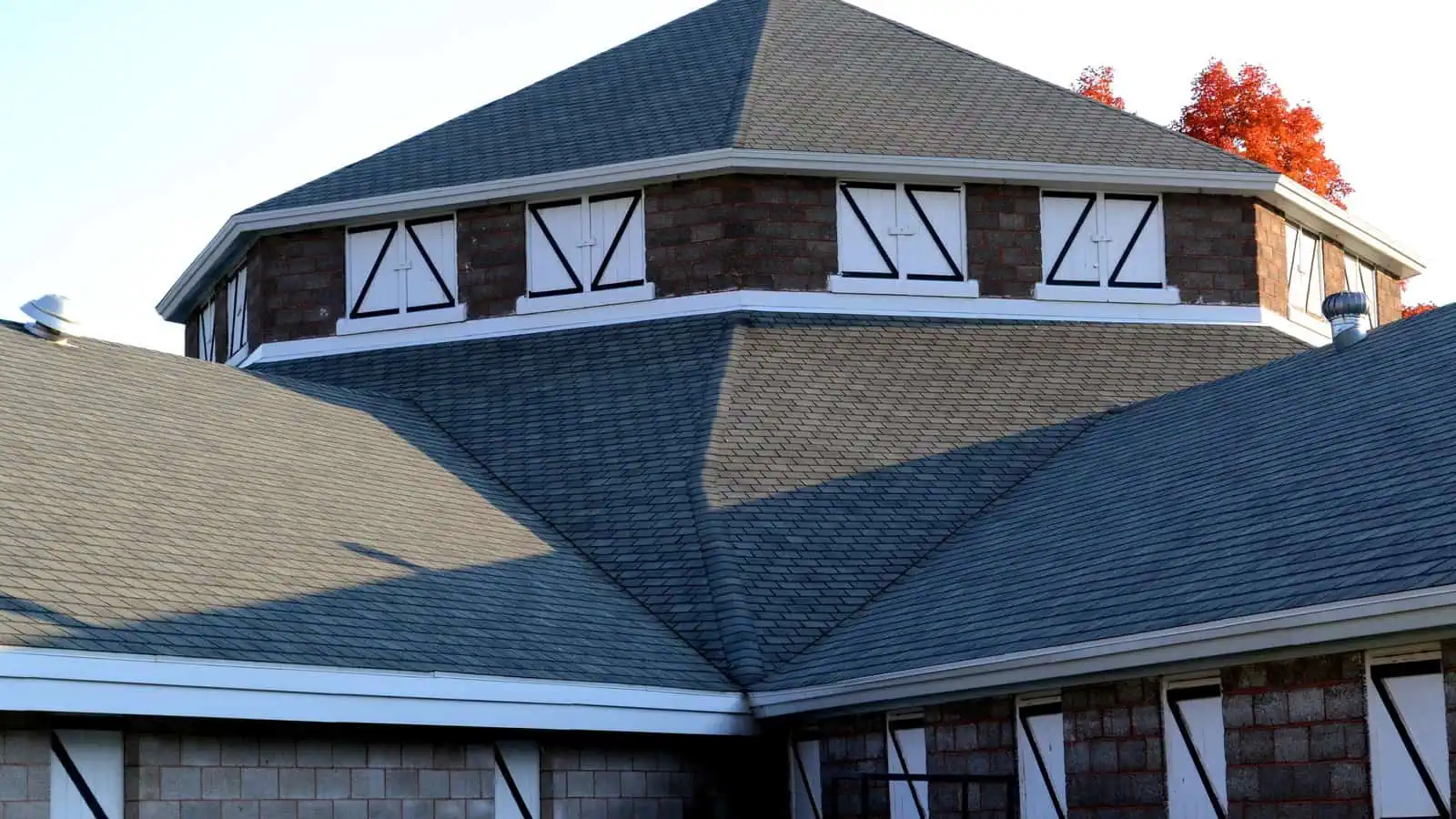 The upper story of an eight-sided concrete-block building with a steep pointed roof, white-trimmed shuttered windows on each face, and the roofs of lower wings converging beneath it, a maple in autumn color at the corner.