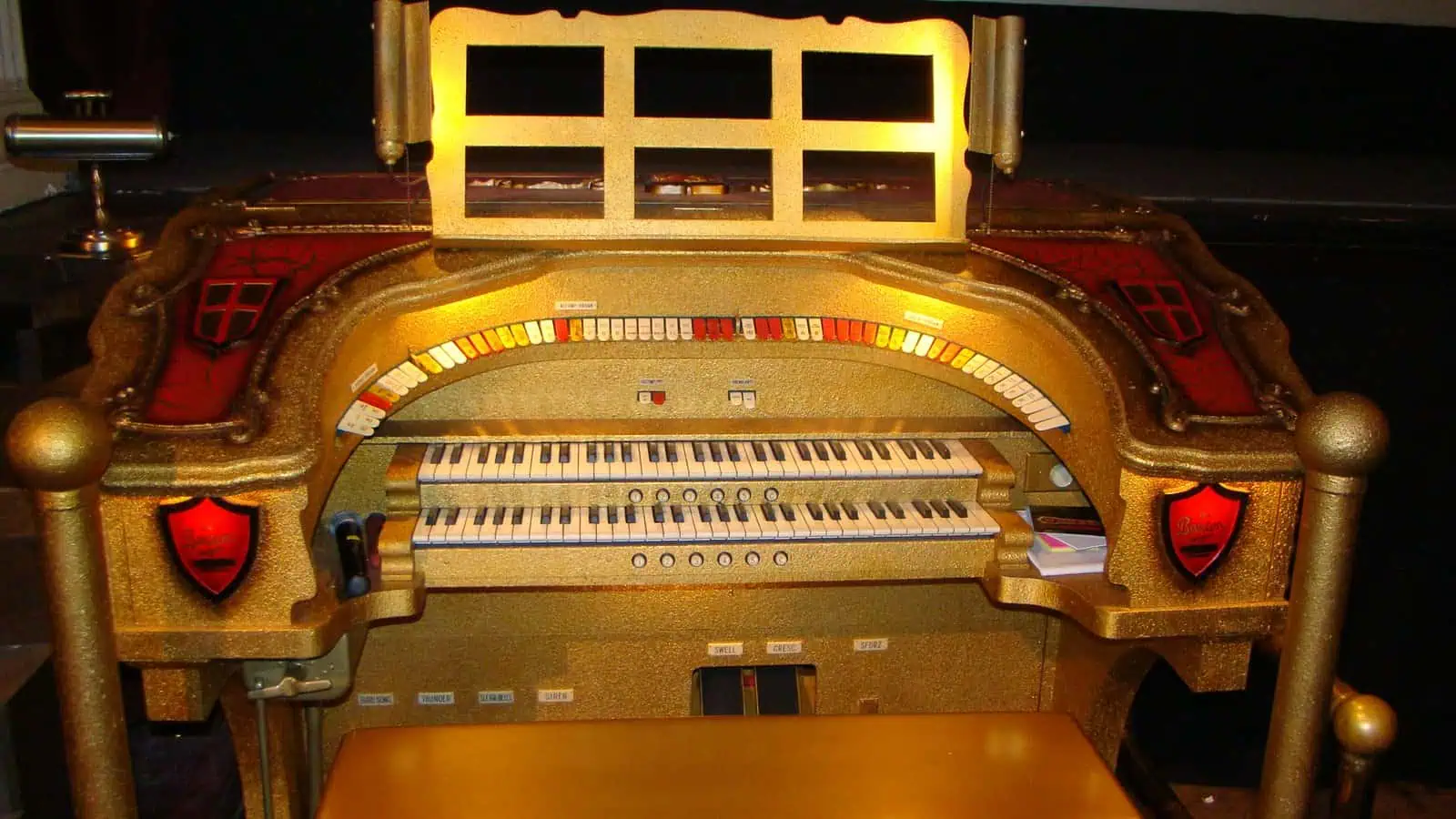 A gilded theatre pipe organ console with two keyboards, an arc of coloured stop tabs above them and a matching gold bench, standing in a dim auditorium.