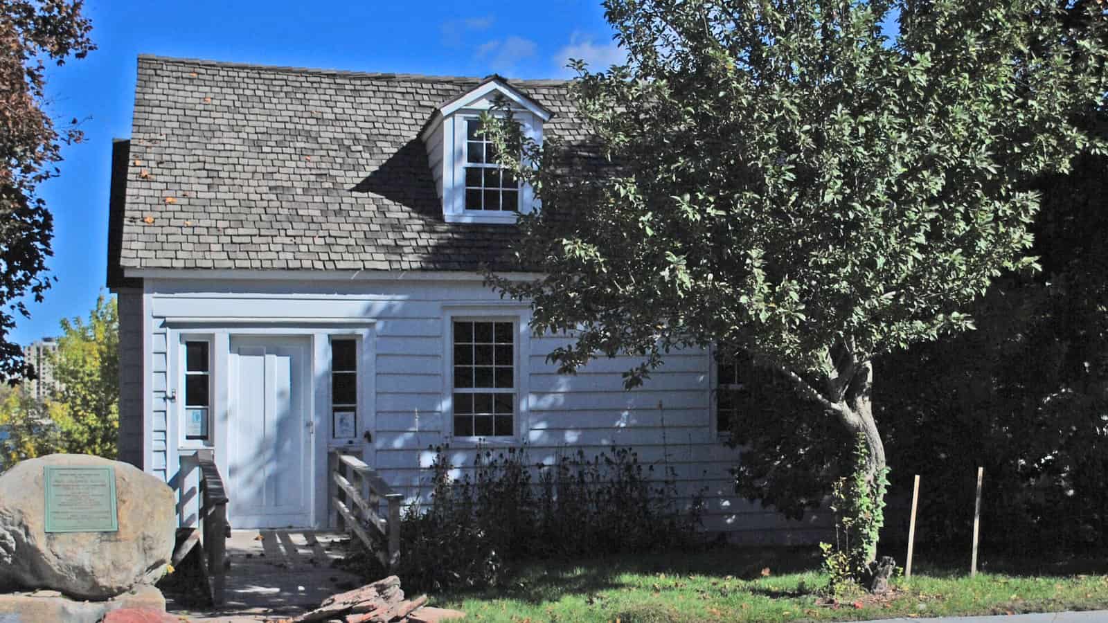 A small white clapboard house with a shingled roof and single dormer window, a plaque mounted on a boulder in the front yard, and a large tree overhanging the right side.