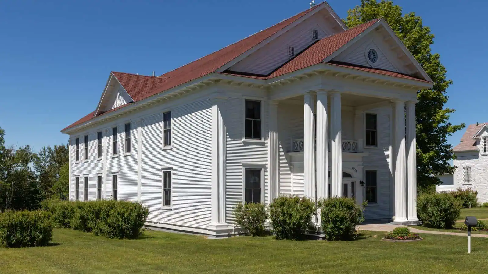 A two-story white clapboard courthouse with a red roof and a tall pedimented portico on six white columns, on a mown lawn under a clear blue sky, a smaller white building visible at the right.