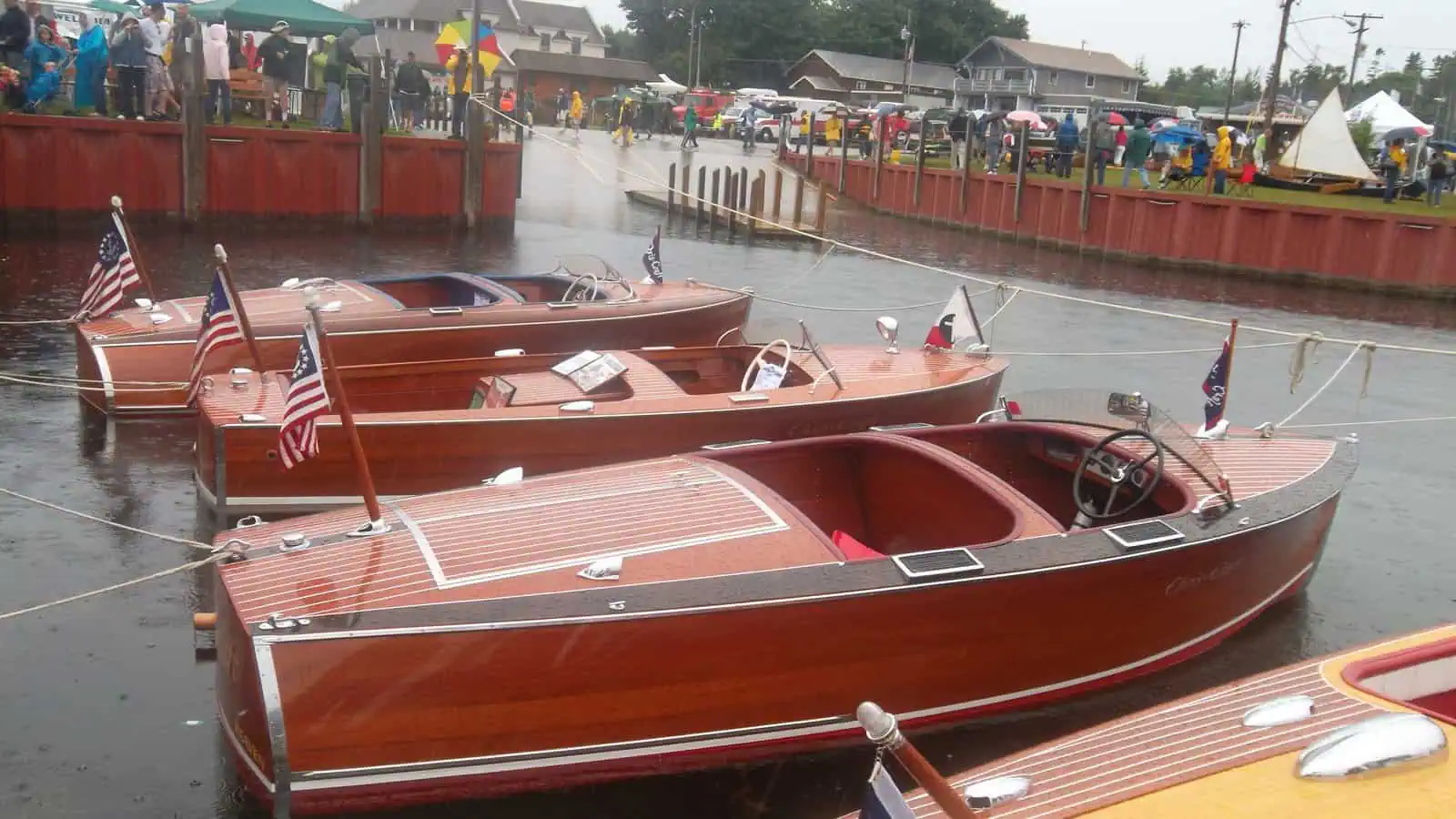 Four varnished mahogany runabouts, each flying a small American flag, moored side by side at a red-railed dock at the Les Cheneaux Antique Wooden Boat Show, with a crowd in rain jackets along the dock behind them under a grey sky.