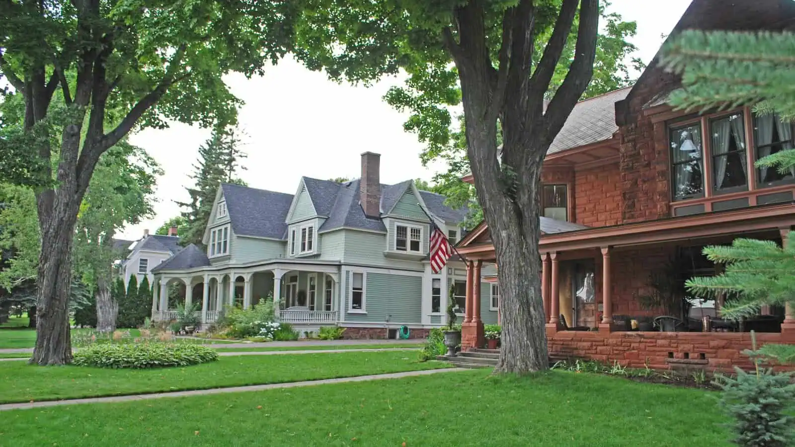 Two large old houses on a leafy Marquette street, a grey-green Queen Anne with a wraparound porch behind and a red sandstone house with a deep front porch in the foreground, big maples over the lawn.