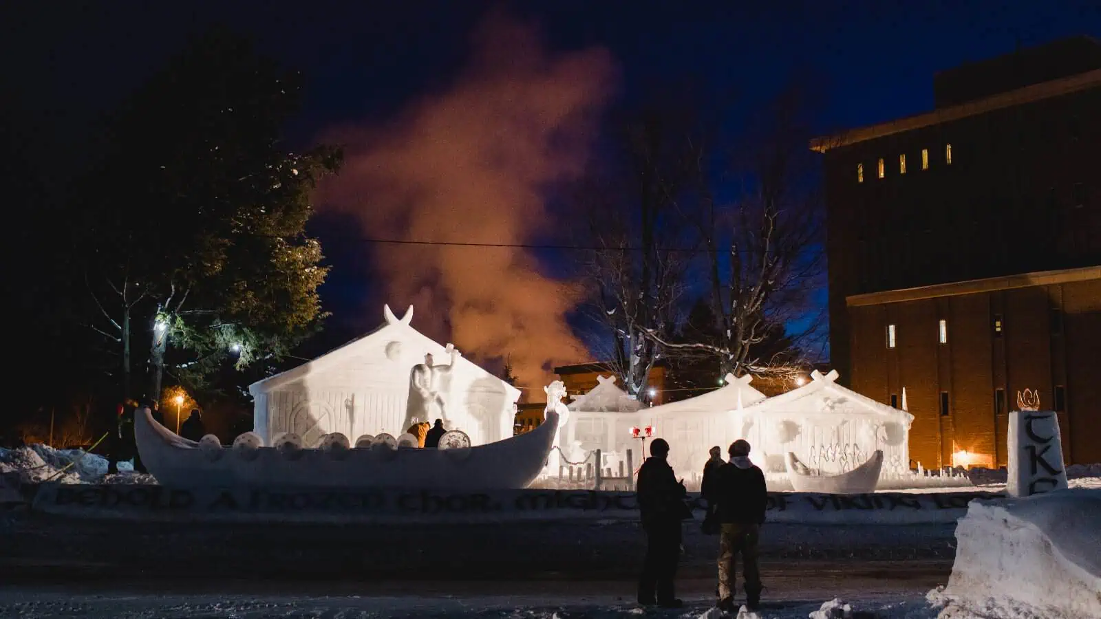 A large carved snow statue of a Viking ship built for Michigan Tech's Winter Carnival, lit at night with onlookers silhouetted beside it.