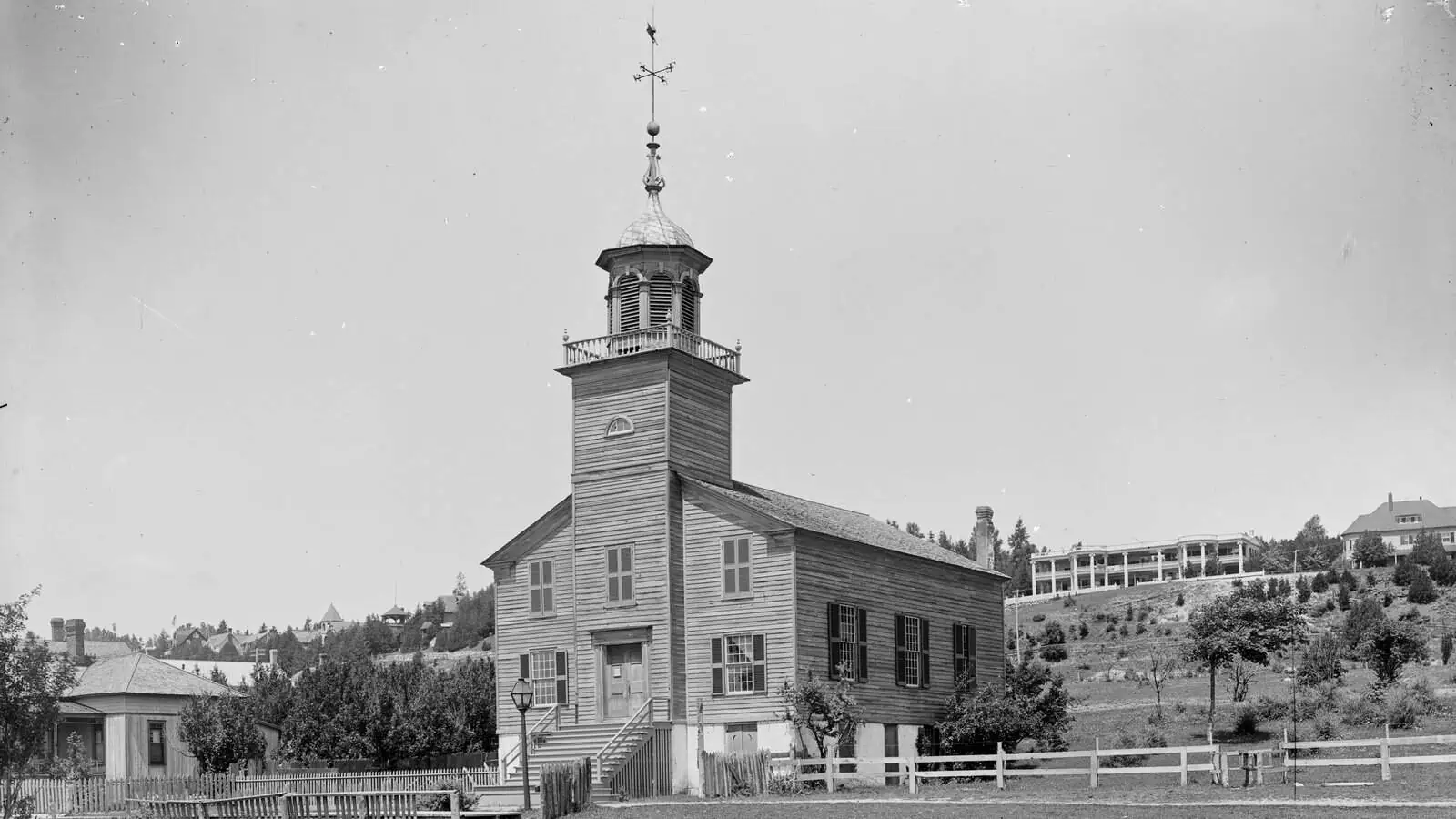 A white clapboard church with a square tower and open belfry topped by a weathervane, standing behind a picket fence on Mackinac Island around 1900, with a wooded hillside and a large hotel building behind it.