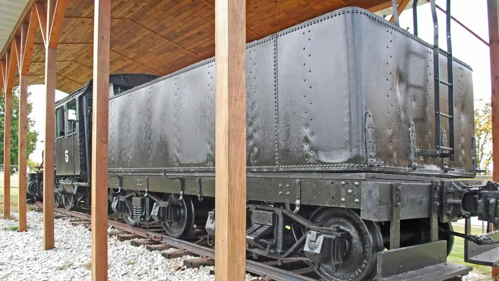 A preserved black steam locomotive and tender standing on a short length of track under an open timber shelter, the number 5 visible on the cab, with gravel underfoot and autumn trees behind.