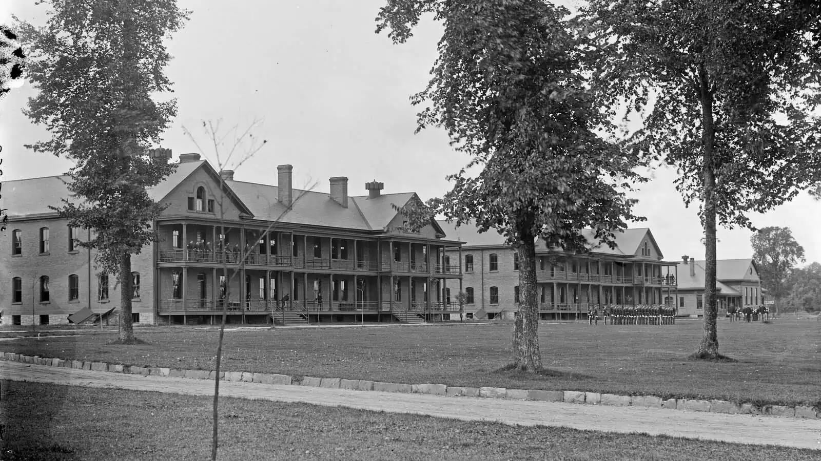 Two long brick barracks with two-story wooden porches face a broad lawn, a company of soldiers standing in formation at the right, large trees in full leaf in the foreground.