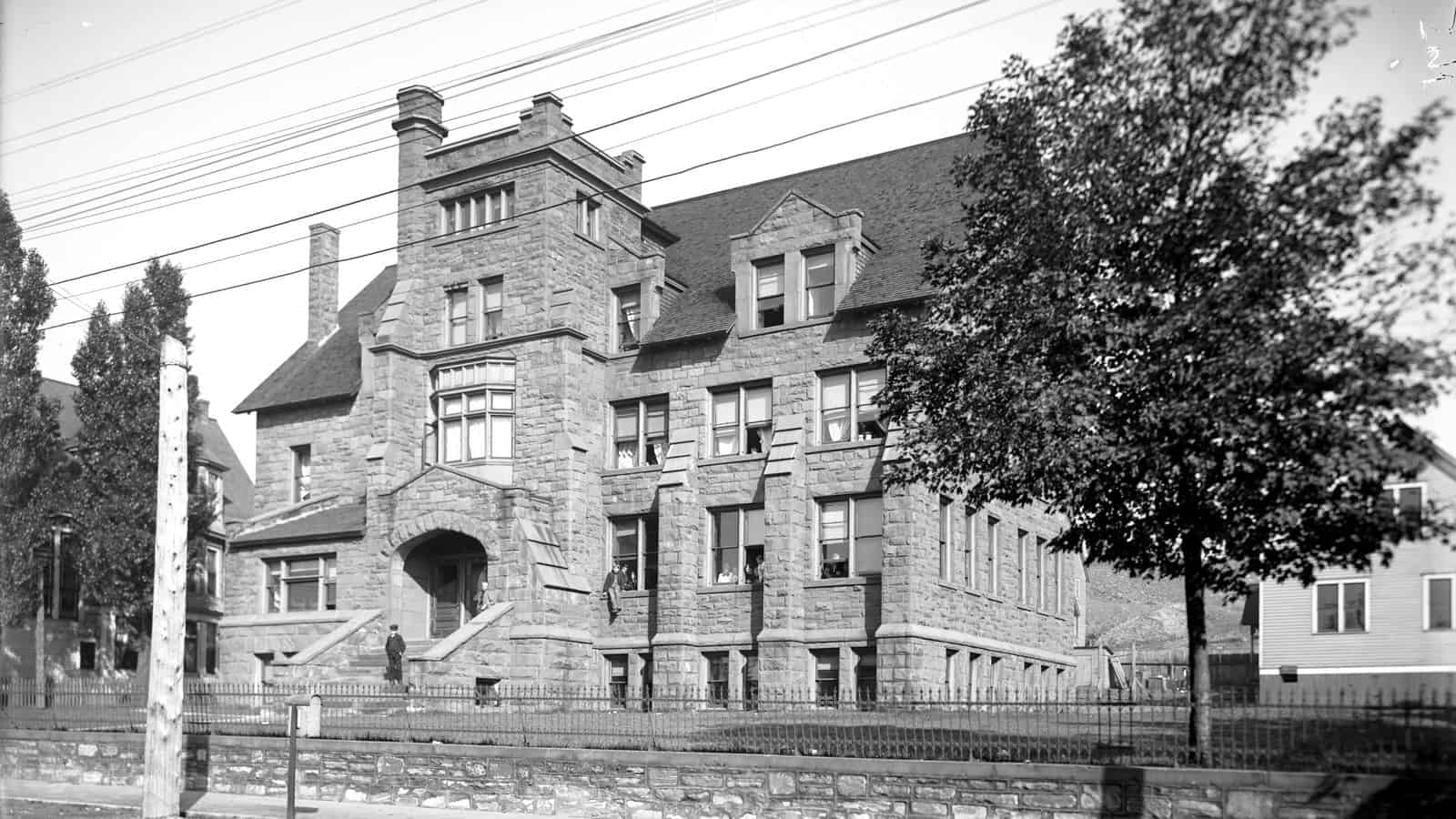 A four-storey rough sandstone college building with a square corner tower, steep gabled roof and an arched stone entrance, behind an iron fence on a stone retaining wall, photographed in the early 1900s with streetcar tracks in the foreground.