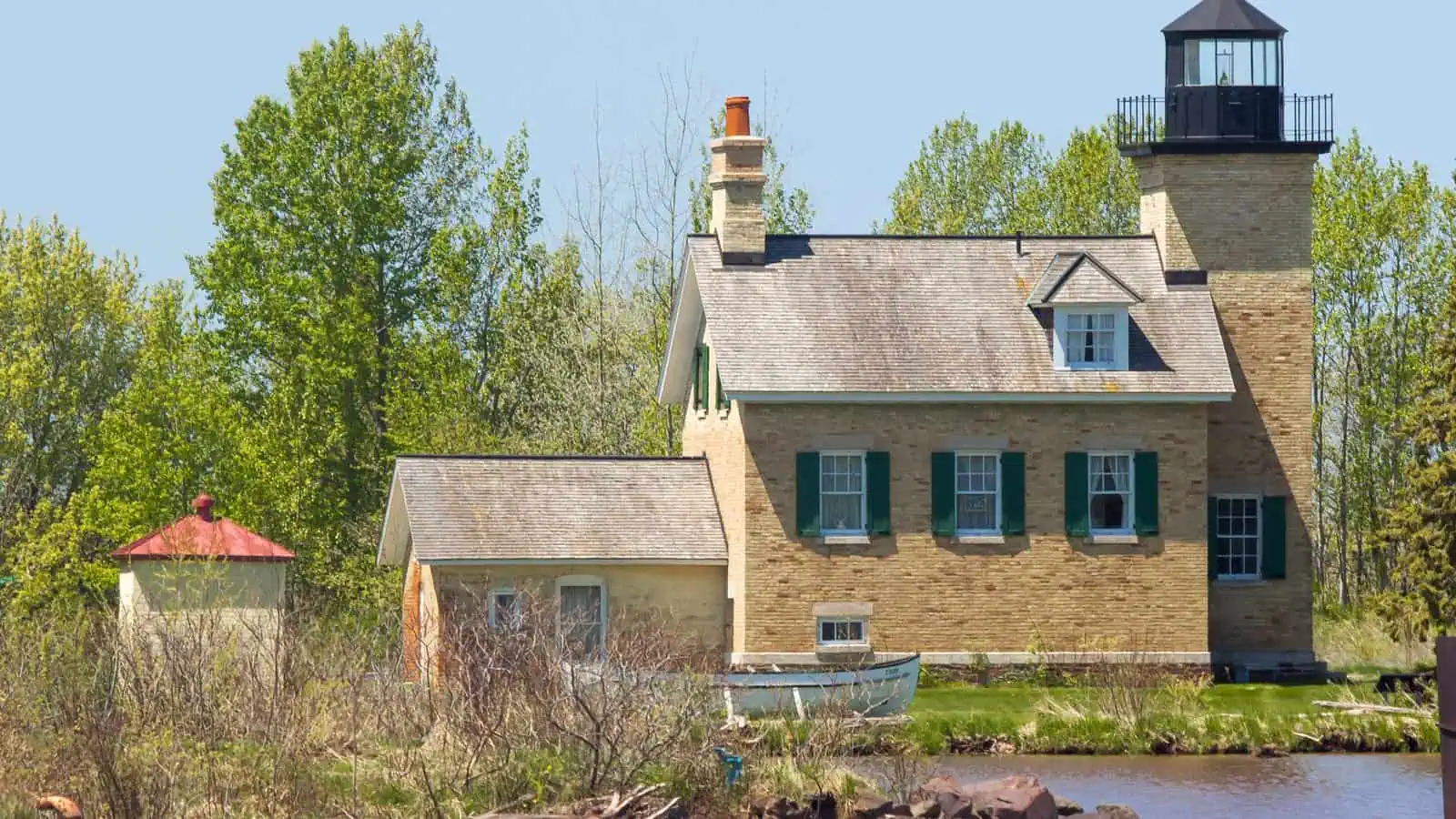 A cream-brick lighthouse with a black lantern rising from the corner of an attached one-and-a-half story keeper's house with green shutters, seen across the river mouth with a small white rowboat on the lawn and a red-roofed oil house at left.