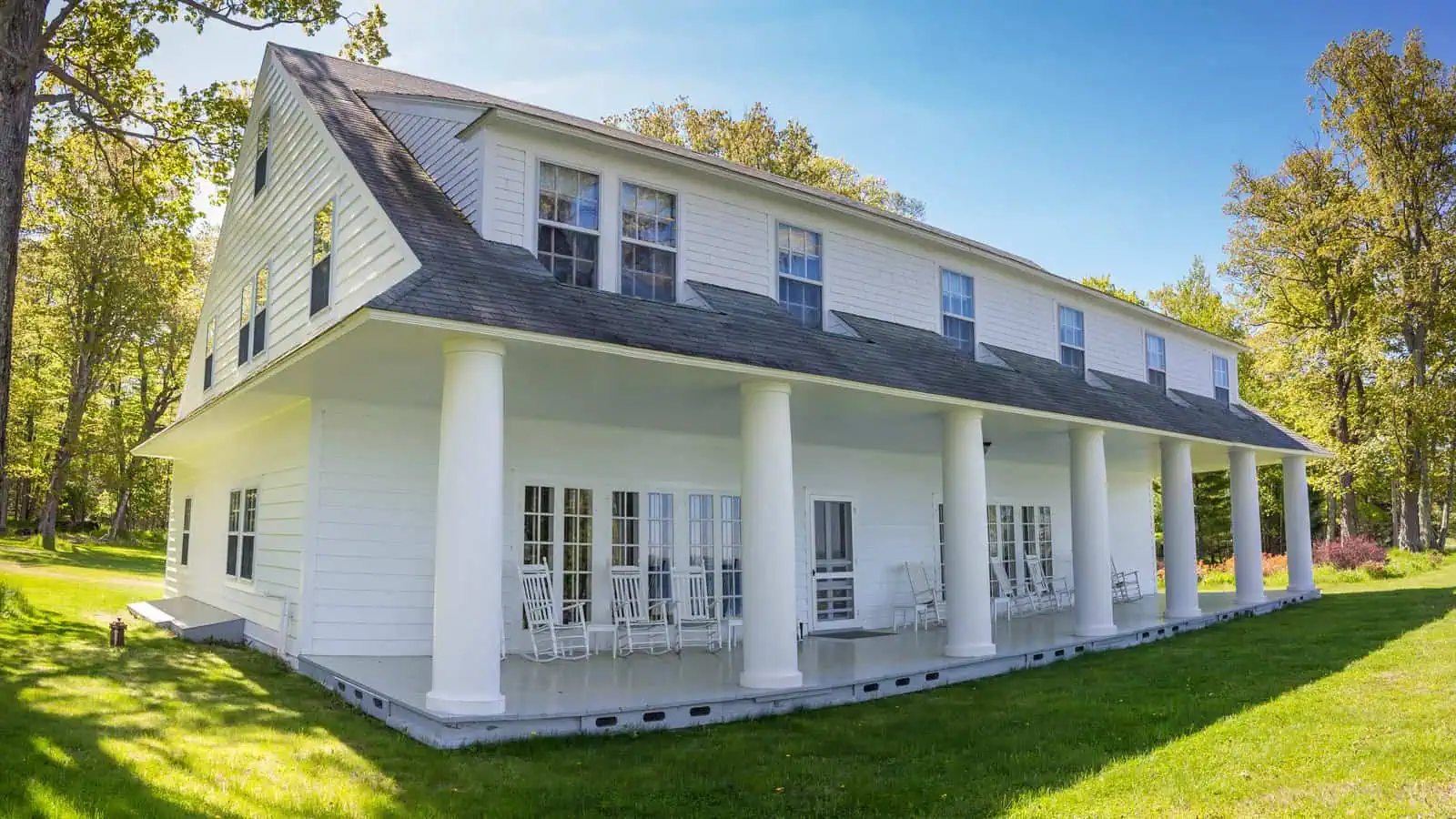 A long two-storey white clapboard house with a grey shingled roof, a row of shed dormers and a deep veranda carried on seven round white pillars, white rocking chairs along the porch, on a mown lawn under trees in bright sun.