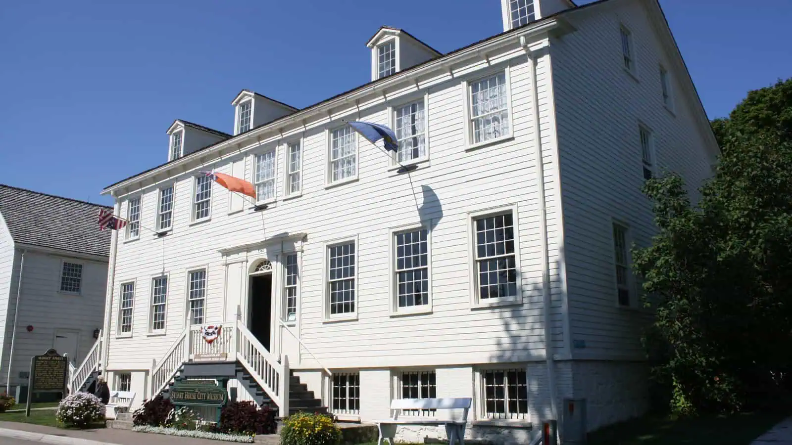 A large white clapboard Federal-style house with a row of dormers, a double front staircase and small flags over the door, a museum sign at the steps, under a clear blue sky.