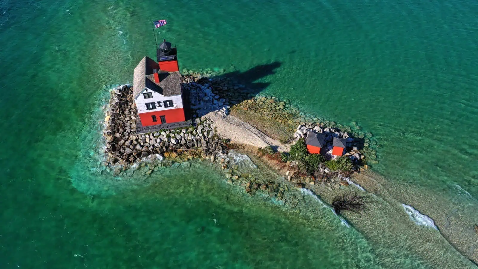 The red and white Round Island Light on its stone point in the turquoise water of the Straits of Mackinac, seen from the air.