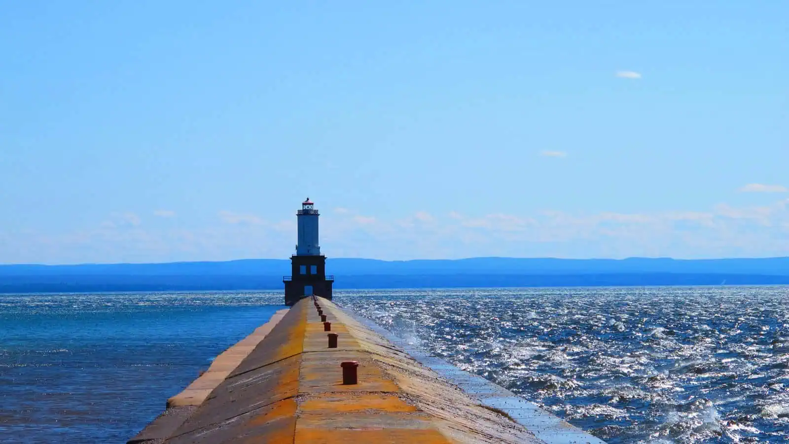 A concrete breakwater runs from the foreground straight out into Lake Superior to a small dark lighthouse, with the far shore low on the horizon under a clear blue sky.