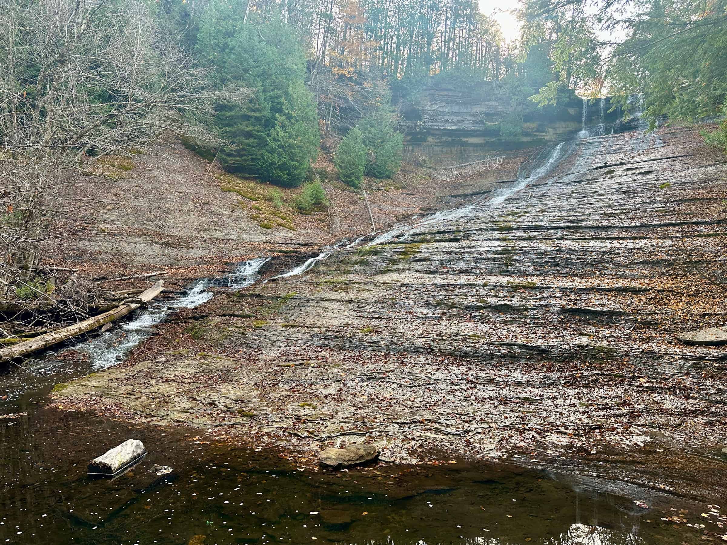 A thin sheet of water slides down a wide, stepped rock face between wooded banks at Laughing Whitefish Falls, with a pool at the bottom. Photo: Seclusive Nature / Flickr (CC BY-SA 4.0).