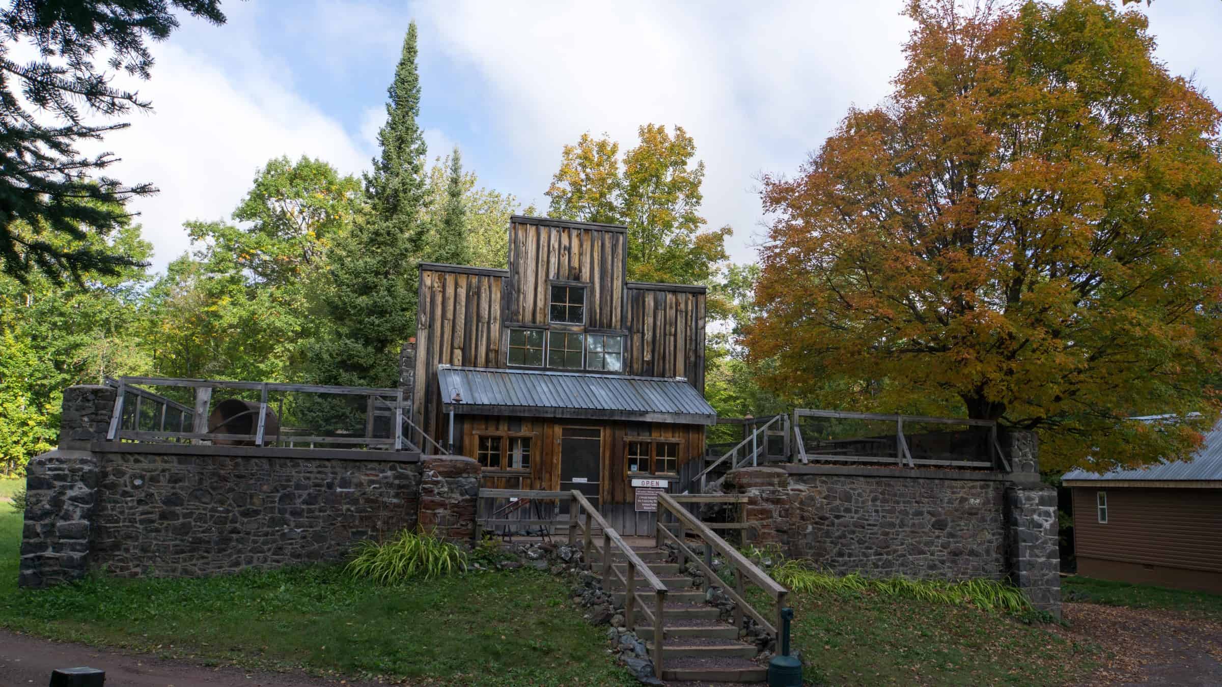 A weathered wooden false-front building with a metal awning sits on a mortared stone wall, with wooden stairs up to the door and an orange maple beside it, at the Adventure Mine in Greenland. Photo: Nathan Miller / National Park Service (Public domain).