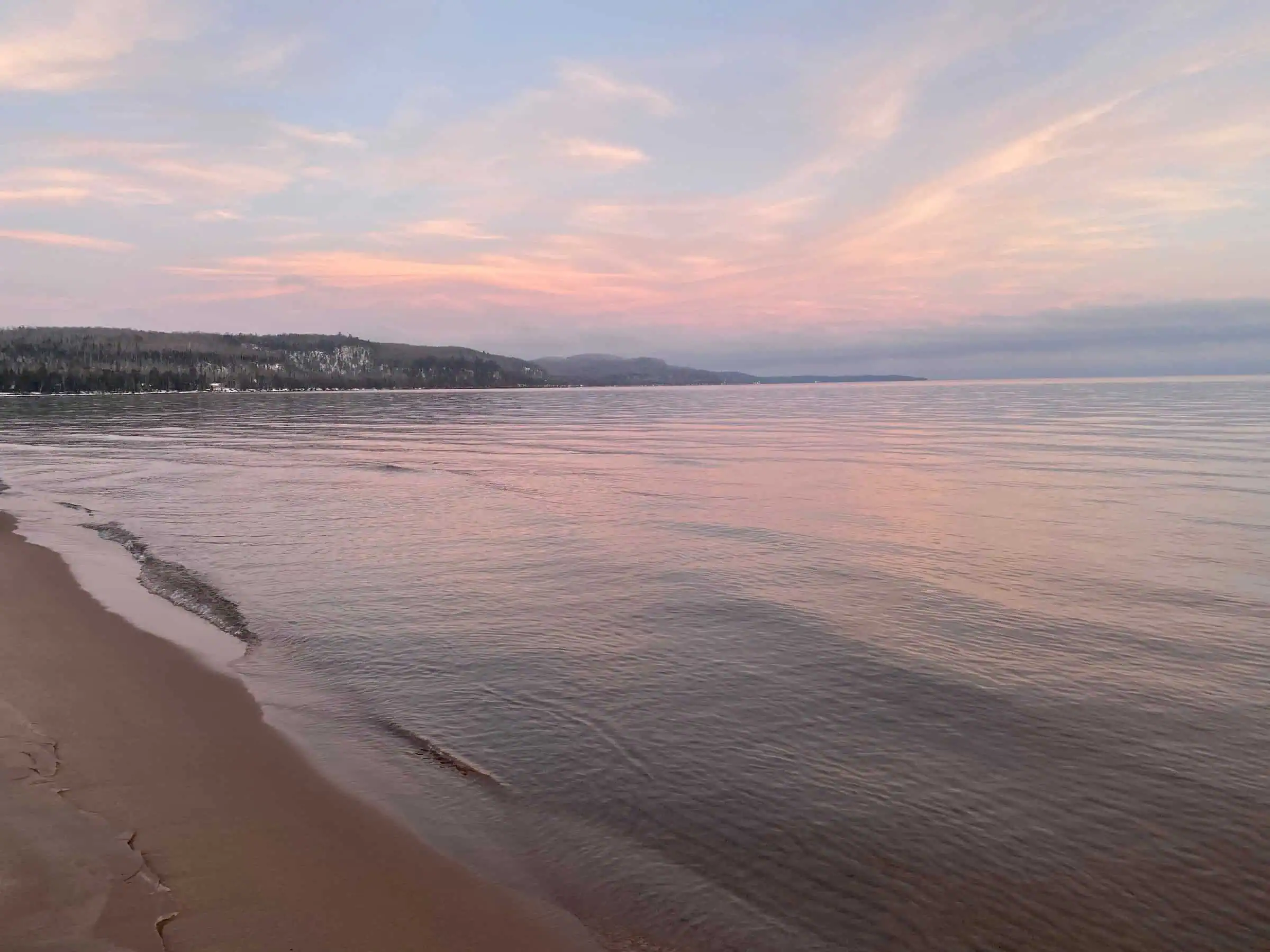 Calm Lake Superior water lapping a sand beach under a pink and blue sky, with a low forested ridge along the far shore. Photo: Fondycardinals / Wikimedia Commons (CC BY-SA 4.0).