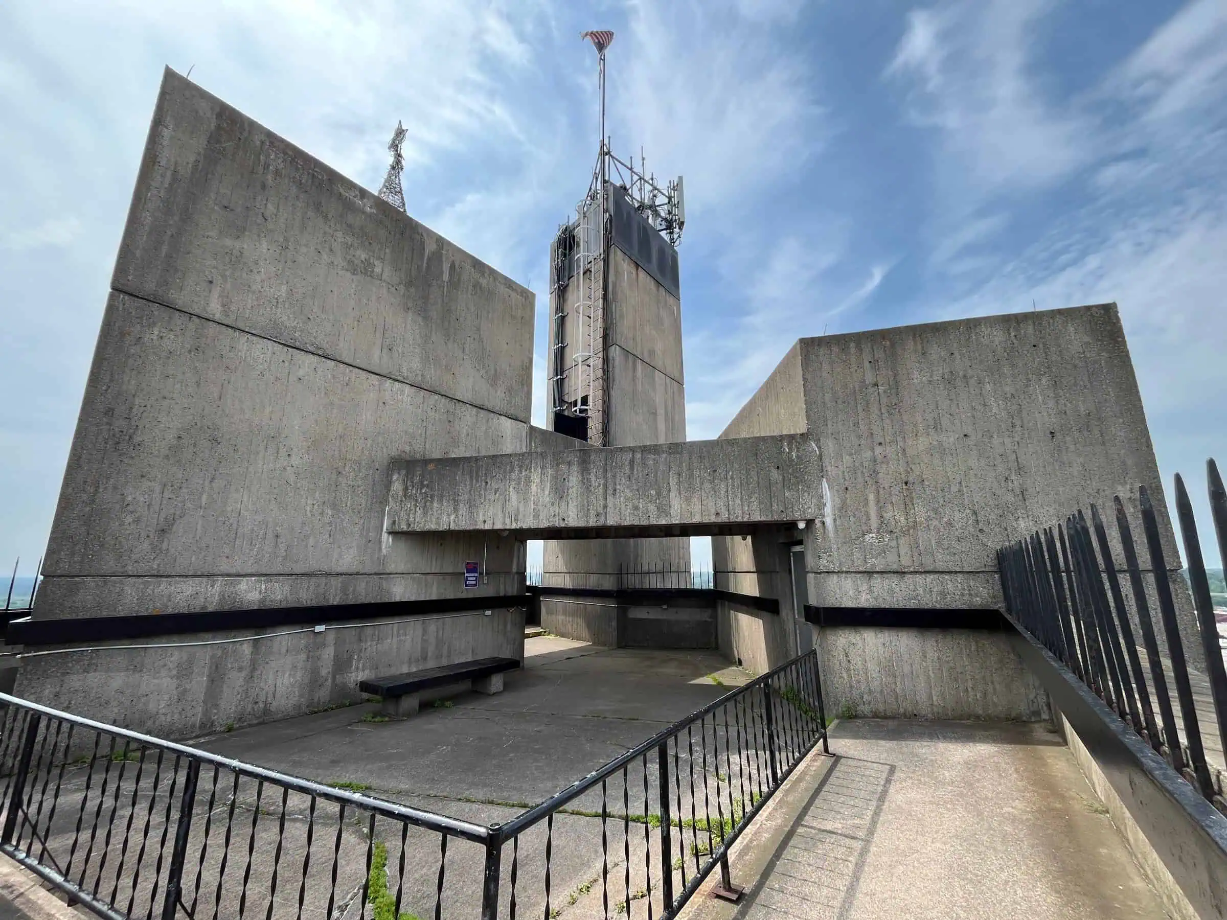 Looking up at a tall, angular concrete tower rising between two slanted concrete wings, with a railing and blue sky with clouds. Photo: Antony-22 / Wikimedia Commons (CC BY-SA 4.0).