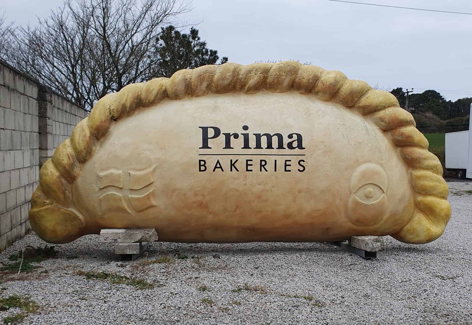 A giant Cornish pasty sculpture bearing the Prima Bakeries name, standing on gravel beside a road in Scorrier, Cornwall. Photo by Mutney, CC BY-SA 4.0.