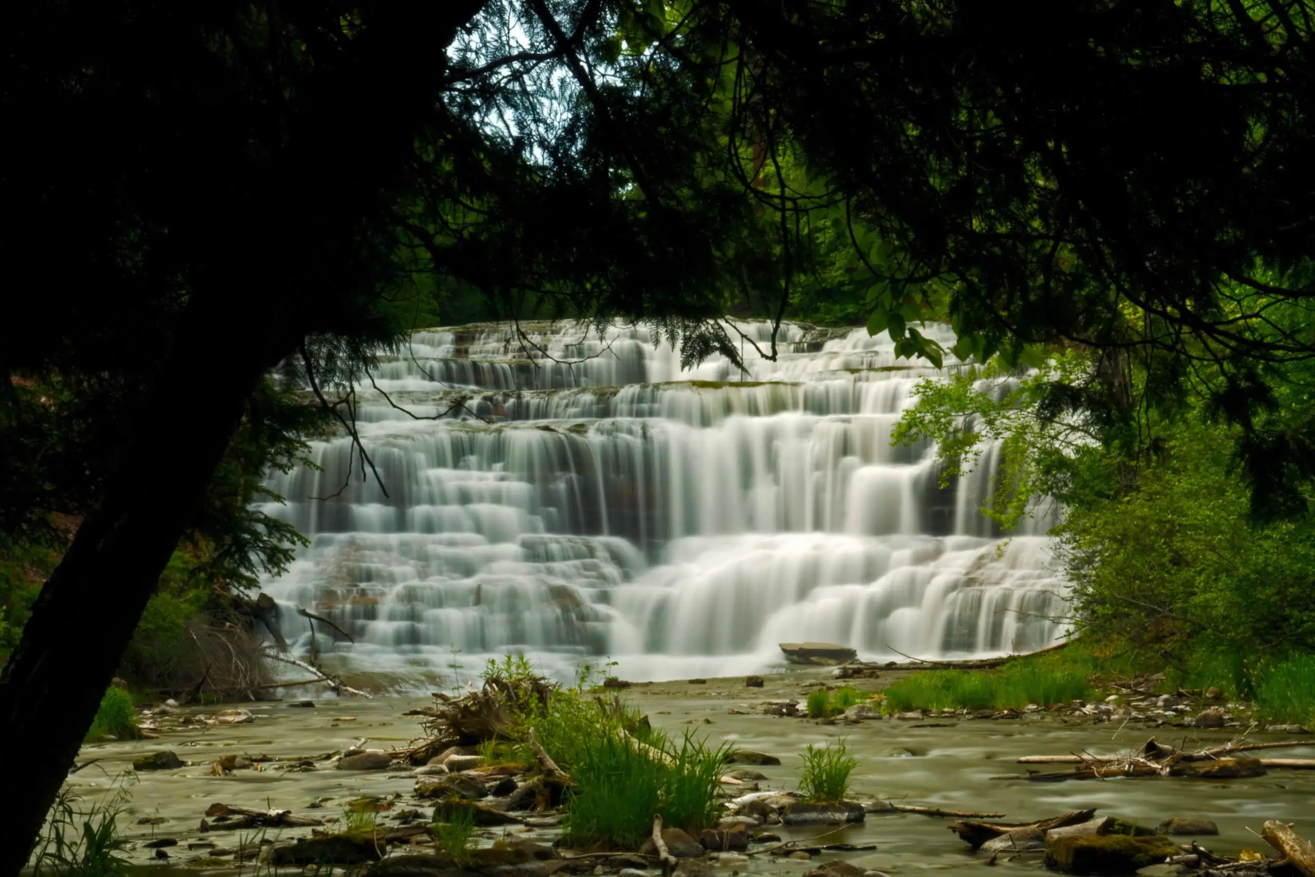 Agate Falls spilling in white steps across a wide sandstone ledge, framed by dark trees in the foreground. Photo: Bobbie Halchishak, U.S. Fish and Wildlife Service / Wikimedia Commons (Public domain).