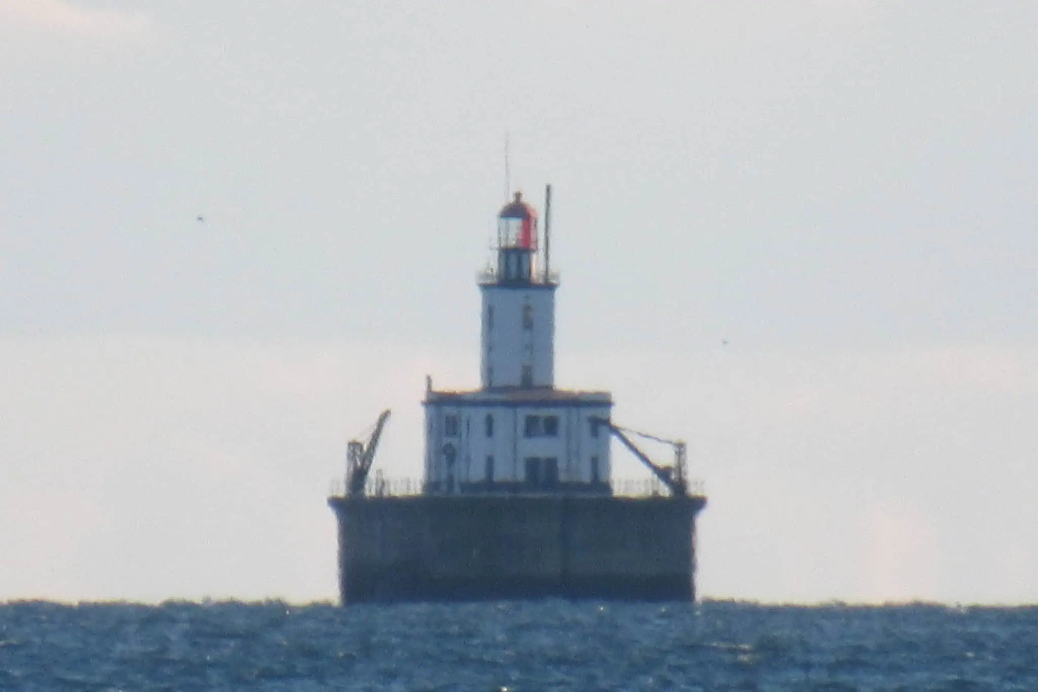 DeTour Reef Light, a white tower with a red lantern on a broad dark base, standing alone in open water. Photo: Notorious4life / Wikimedia Commons (CC0).