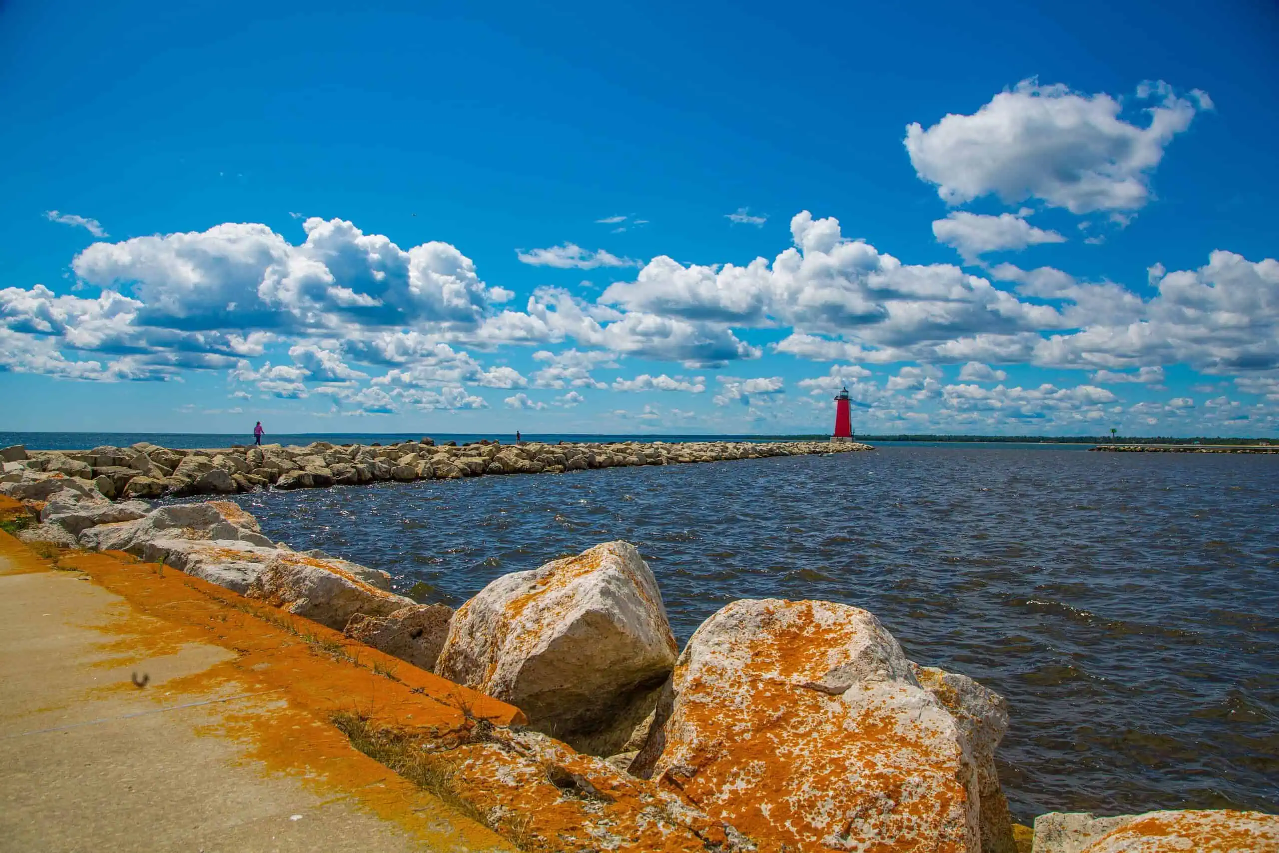 Orange lichen-covered boulders on the Manistique shore, with a concrete breakwater running out to a small red lighthouse on Lake Michigan. Photo: Nuggehalli2015 / Wikimedia Commons (CC BY-SA 4.0).