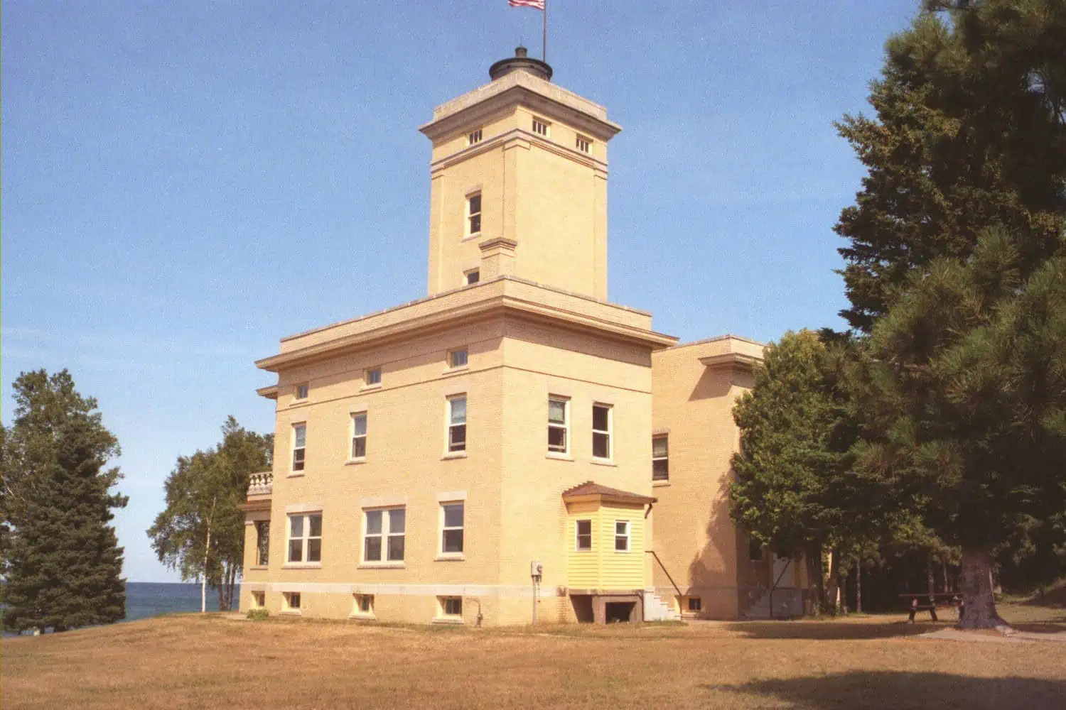 Sand Hills Lighthouse, a buff brick building with a square central tower, on a lawn ringed by trees, photographed in August 2006. Photo: Chris Light / Wikimedia Commons (CC BY-SA 4.0).